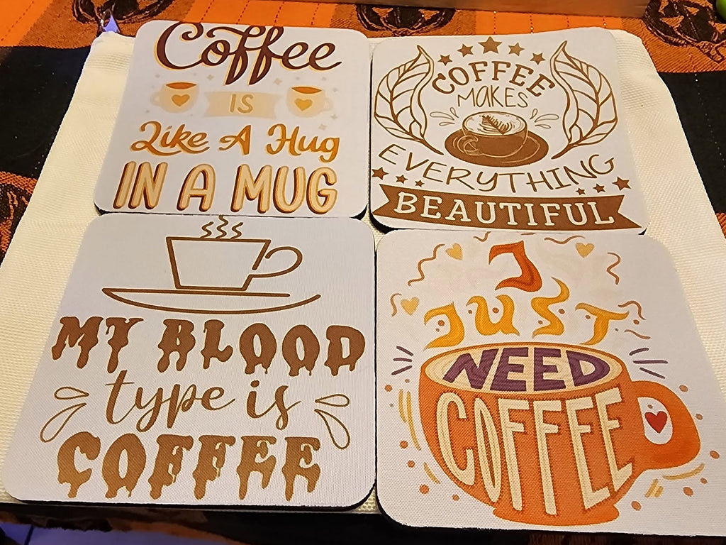 Four square coasters with coffee-themed quotes and illustrations, arranged on a dark patterned tablecloth. The coasters are white with brown and orange graphics and text including 'Coffee is like a hug in a mug', 'Coffee makes everything beautiful', 'My blood type is coffee', and 'I just need coffee'.