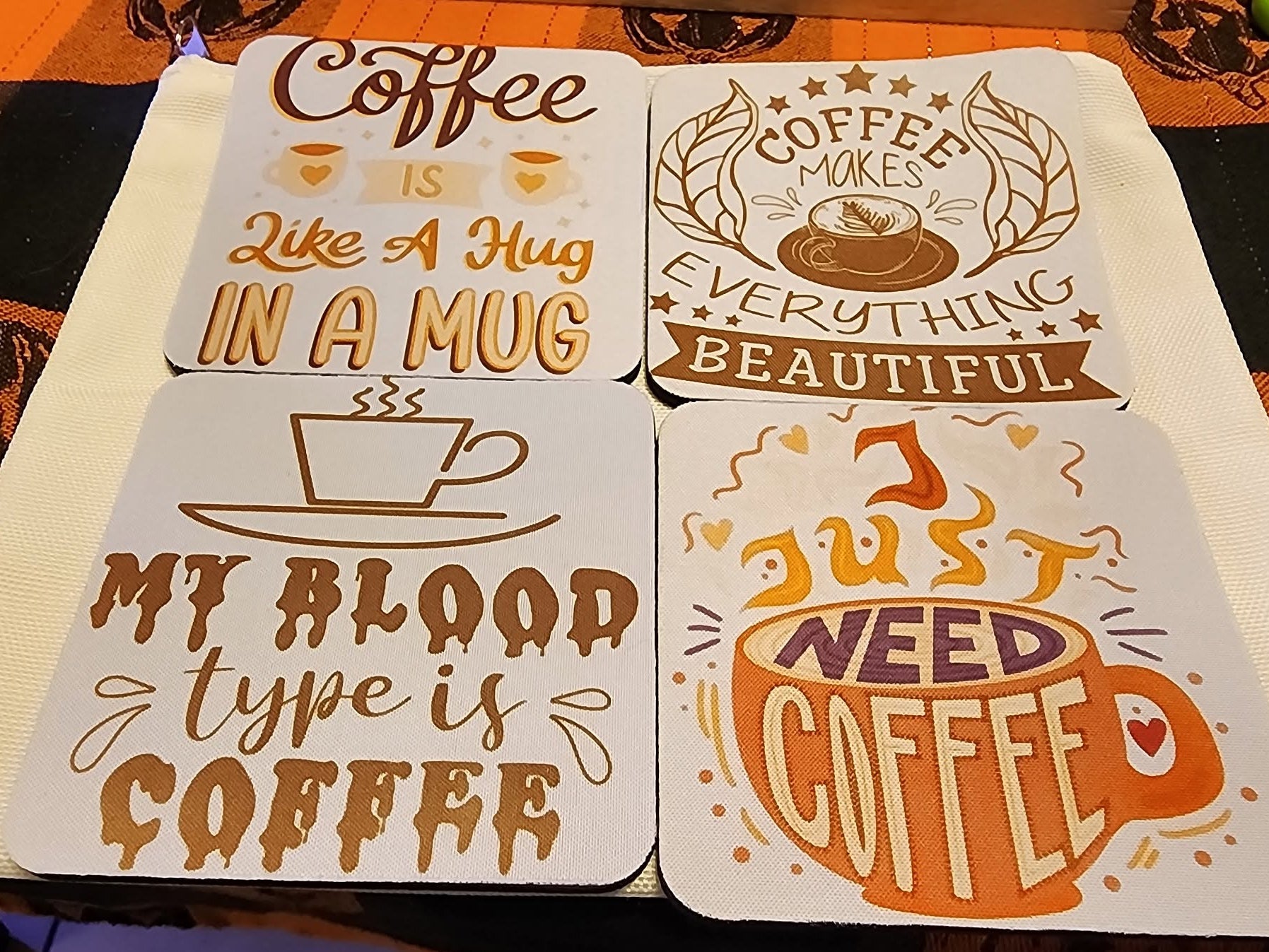 Four square coasters with coffee-themed quotes and illustrations, arranged on a dark patterned tablecloth. The coasters are white with brown and orange graphics and text including 'Coffee is like a hug in a mug', 'Coffee makes everything beautiful', 'My blood type is coffee', and 'I just need coffee'.