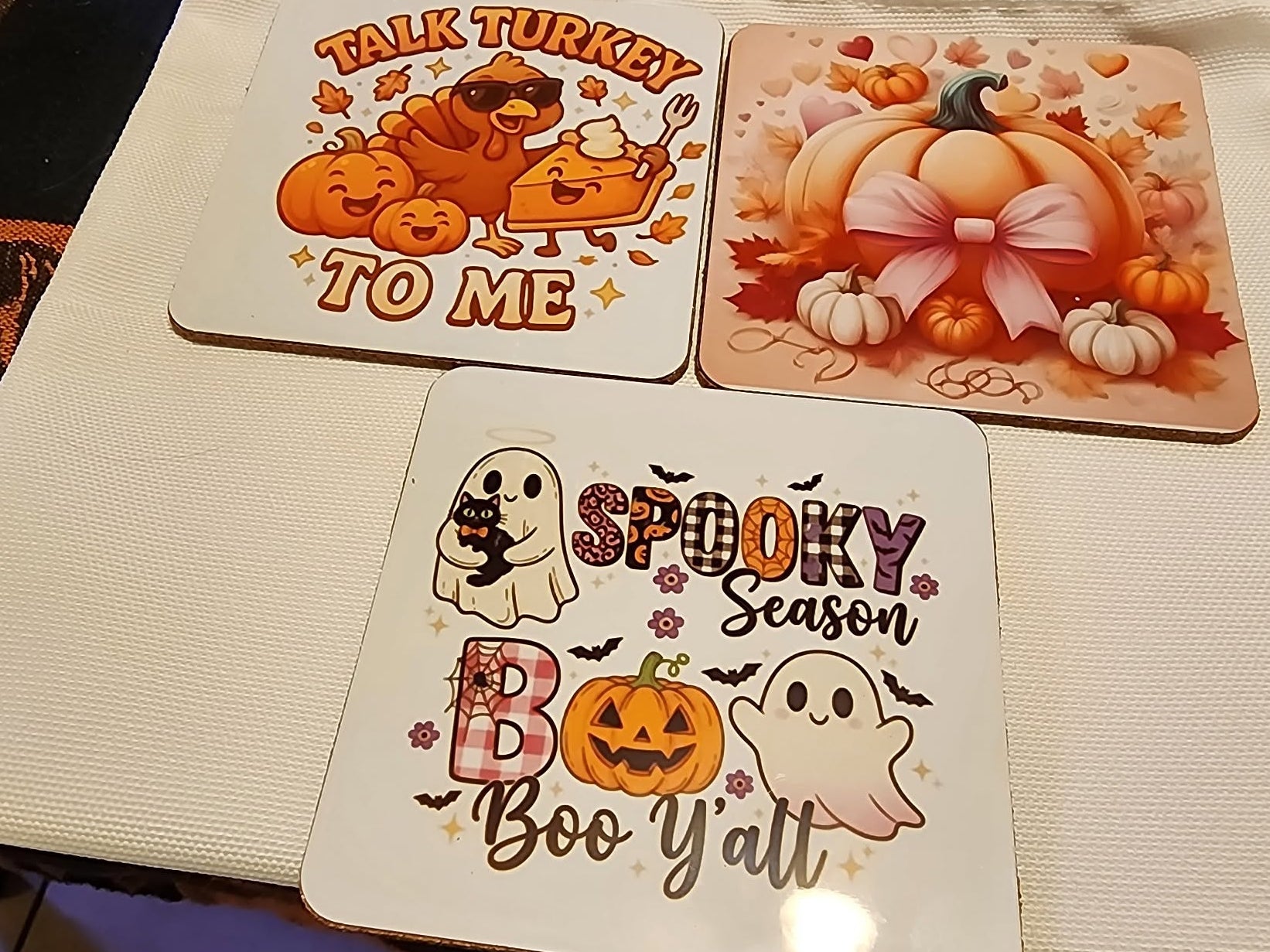 Three square Halloween-themed coasters displayed on a white textured fabric surface. The coasters feature autumn and spooky illustrations: one with pumpkins and the text 'TALK TURKEY TO ME', one with pumpkins and a bow reading 'BOY', and one with ghosts, pumpkins, and the text 'SPOOKY Season Boo Y'all'.